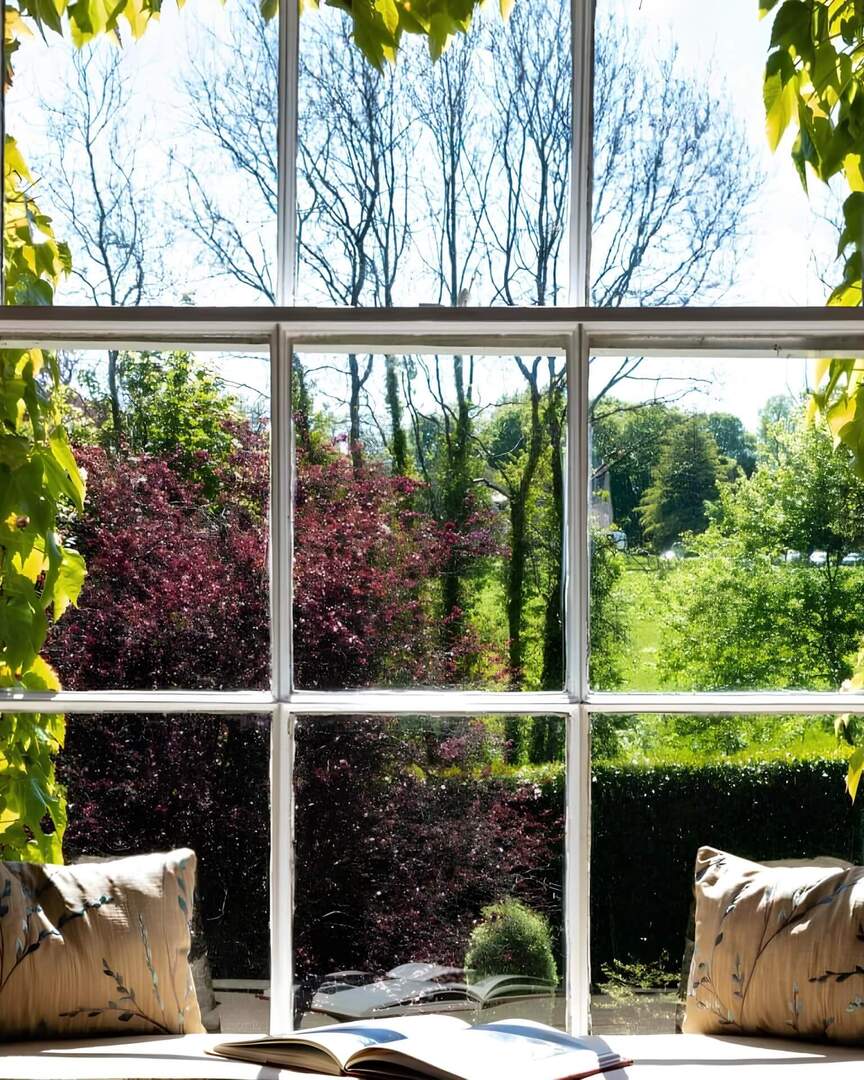 Coin fenêtre ensoleillé avec coussins et livre ouvert, vue sur lierre, arbustes en fleurs et jardin verdoyant