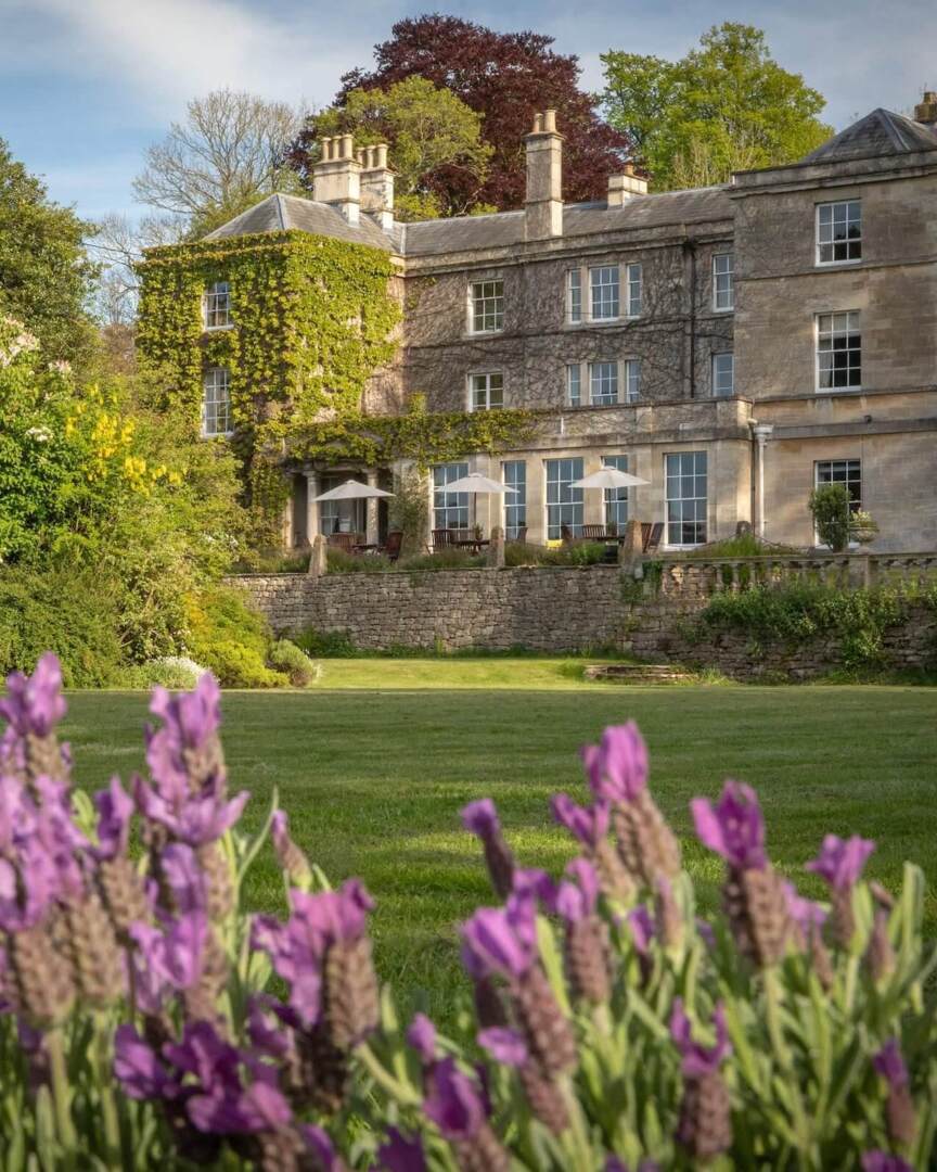 Historic stone hotel with ivy-covered walls, outdoor terrace with umbrellas, surrounded by lush gardens and blooming purple flowers