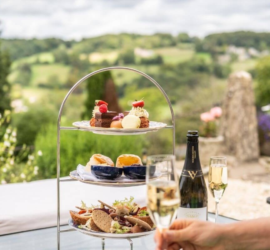 Three-tier afternoon tea stand with pastries, scones, and sandwiches on an outdoor table with champagne glasses and countryside views
