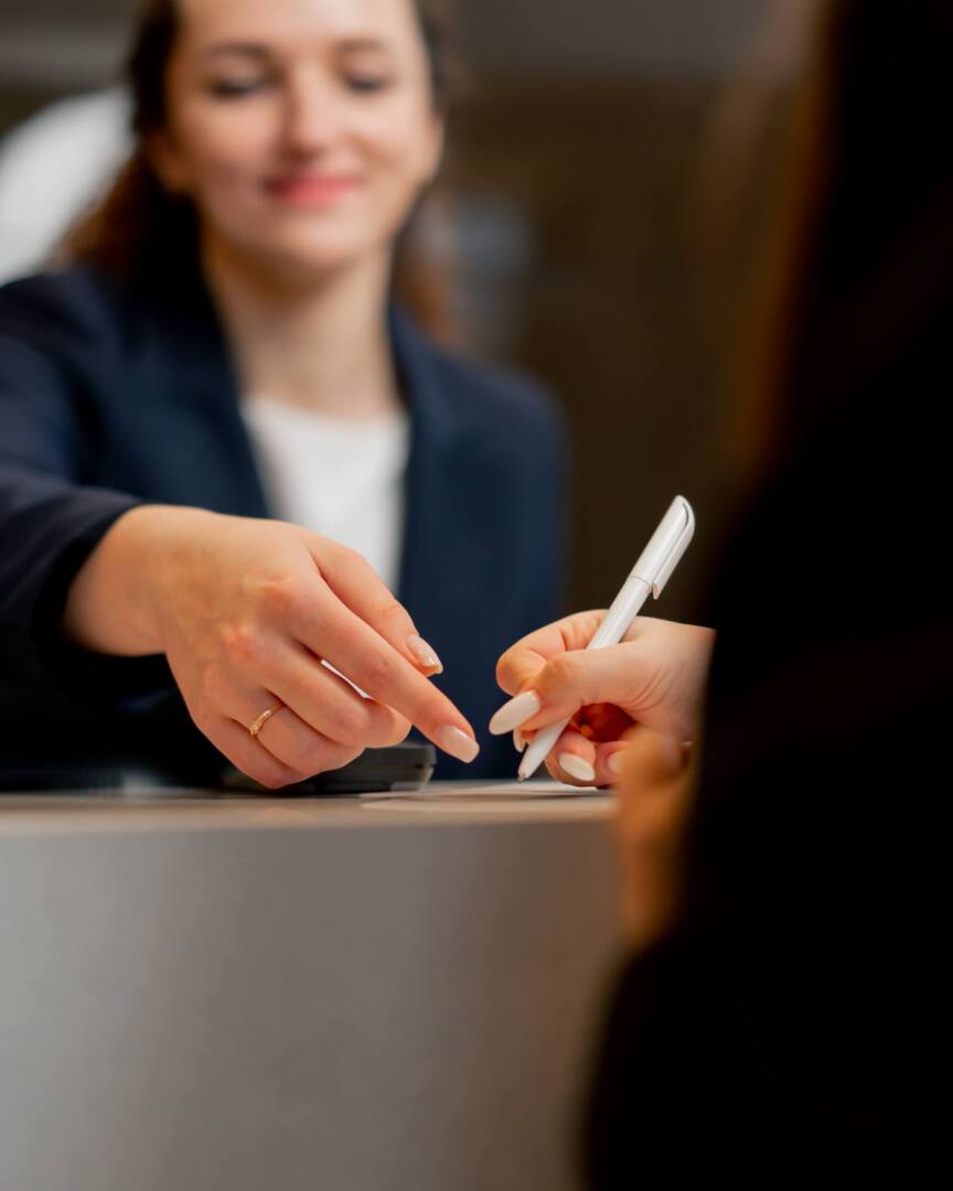 Hotel receptionist assisting guest with check-in, smiling and pointing at the document to sign at the front desk