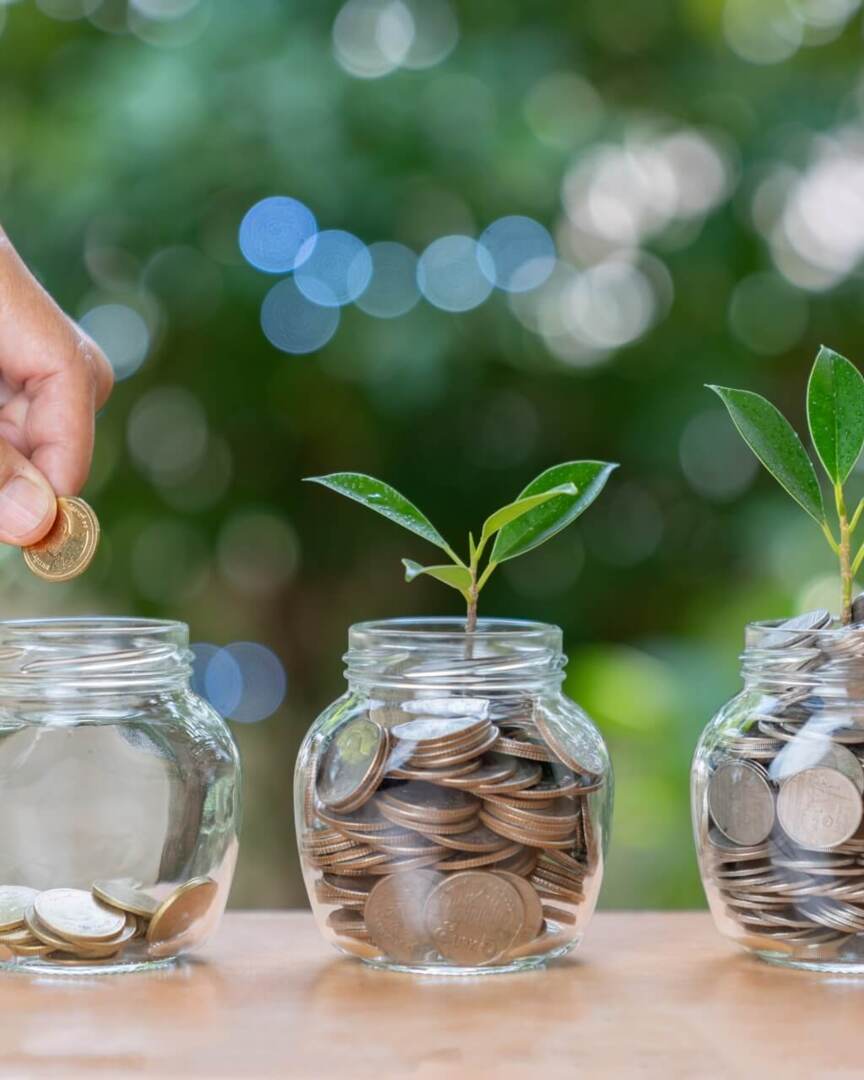 Hand placing coin in jar with coins growing into plants, symbolizing savings and growth against blurred green background