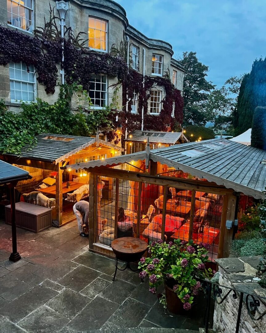 Historic hotel courtyard with ivy-covered stone building, cozy outdoor seating in wooden pavilions lit by string lights at dusk