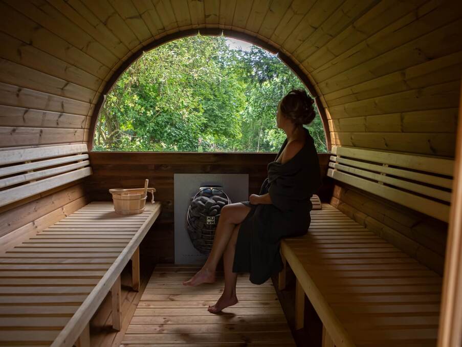 Sauna tonneau en bois, femme assise en peignoir près du poêle, seau pour l'eau, fenêtre arrondie sur un jardin verdoyant