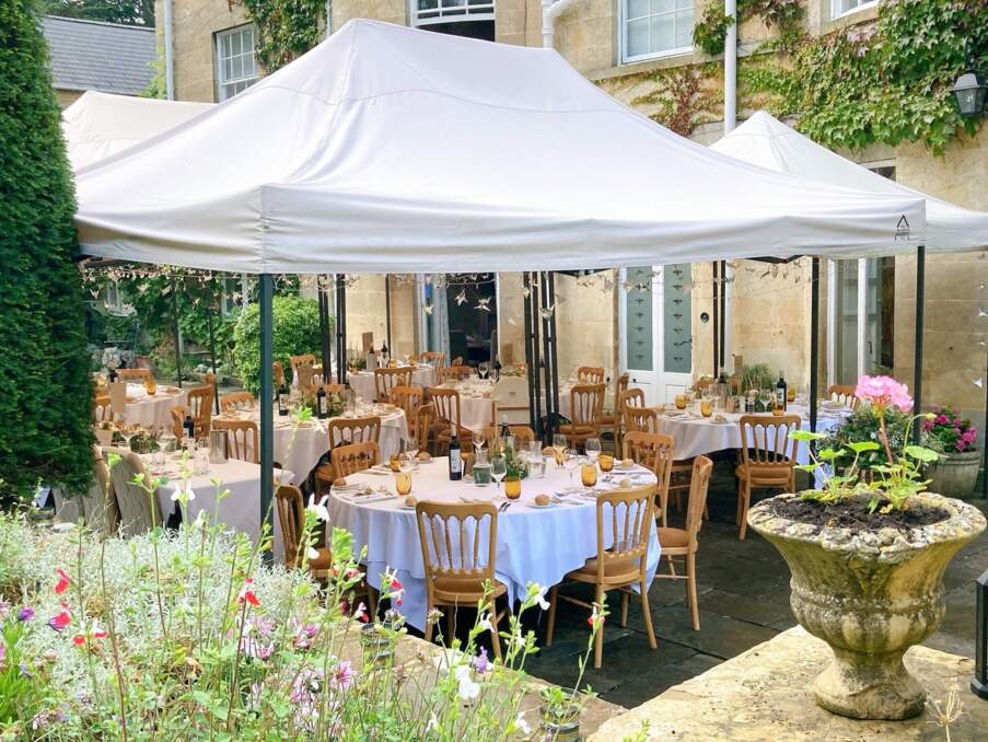 Outdoor hotel dining area under white canopy with round tables, wooden chairs, floral centerpieces, and warm glassware set for guests
