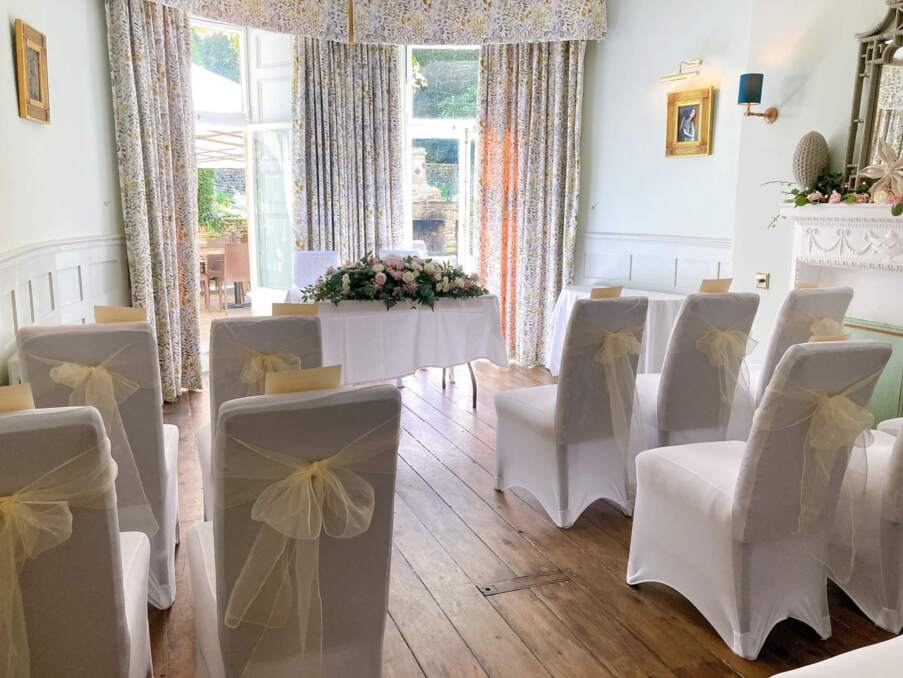 Bright wedding ceremony room with white chairs draped in sheer bows, floral centerpiece, wooden floors, and floral curtains overlooking garden.