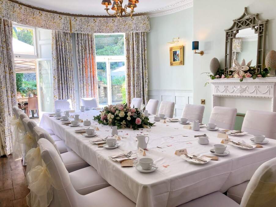 Elegant dining room set for event with white tablecloth, floral centerpiece, cushioned chairs, and natural light from large windows