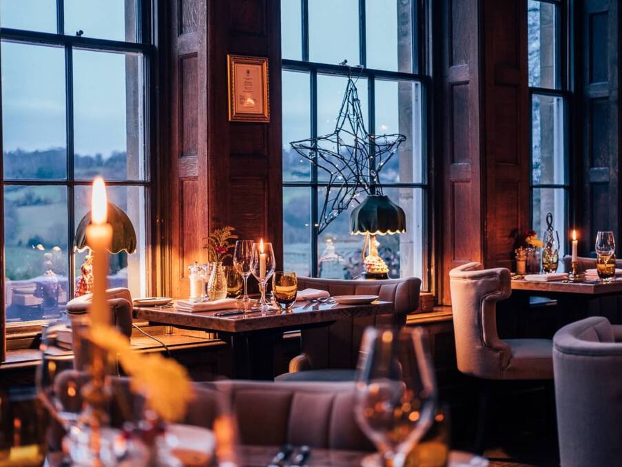 Cozy fine dining room with wood paneling, candlelit tables, plush chairs, decorative lamps, and large windows overlooking a countryside view