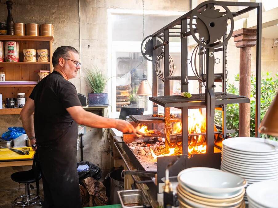 Chef grilling food over an open flame in a rustic kitchen area with shelves of ingredients and stacked white plates nearby