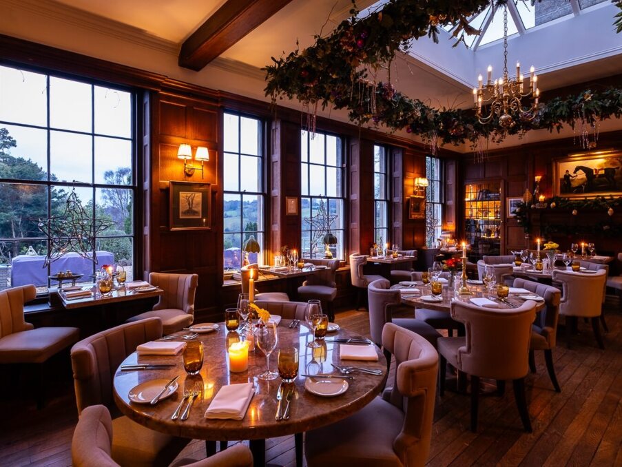 Elegant dining room with plush chairs, candlelit tables, large windows showing scenic countryside, and festive holiday decor with chandelier