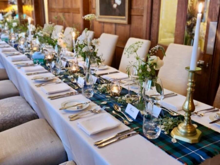 Elegant dining table set with white linens, glassware, fresh floral arrangements, and brass candlesticks in a wood-paneled room