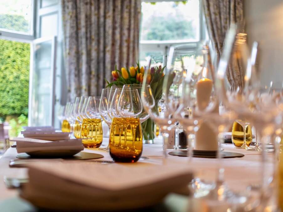 Elegant dining table set with folded napkins, amber glassware, wine glasses, candles, and a floral centerpiece near large windows
