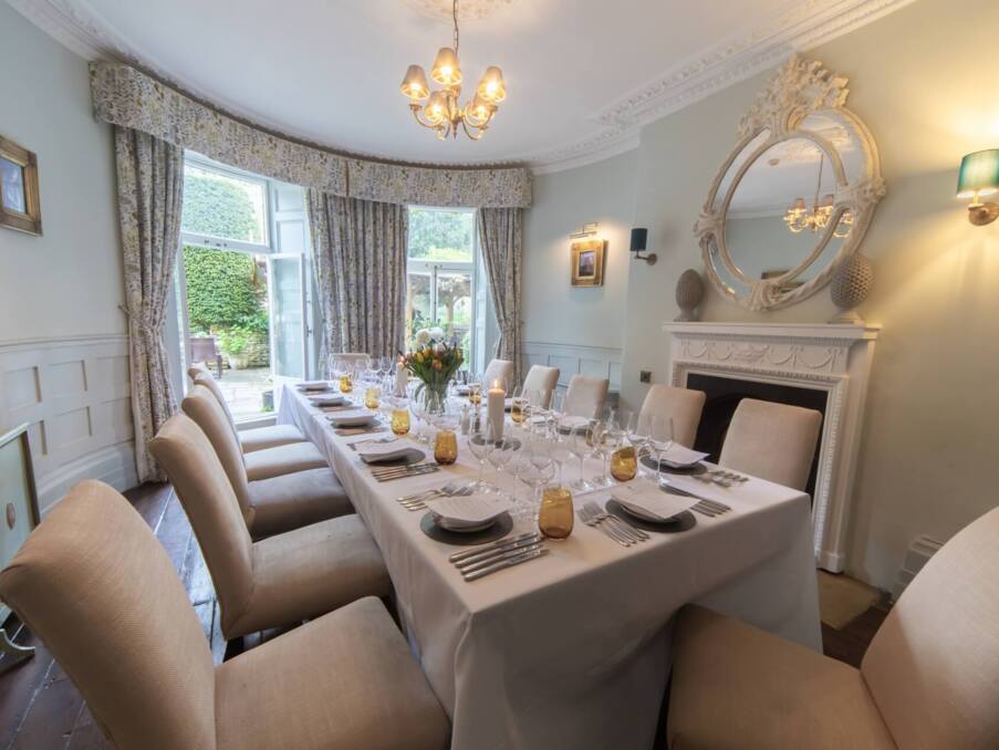 Elegant dining room with ornate fireplace, large round mirror, beige upholstered chairs, floral curtains, and table set for a formal meal