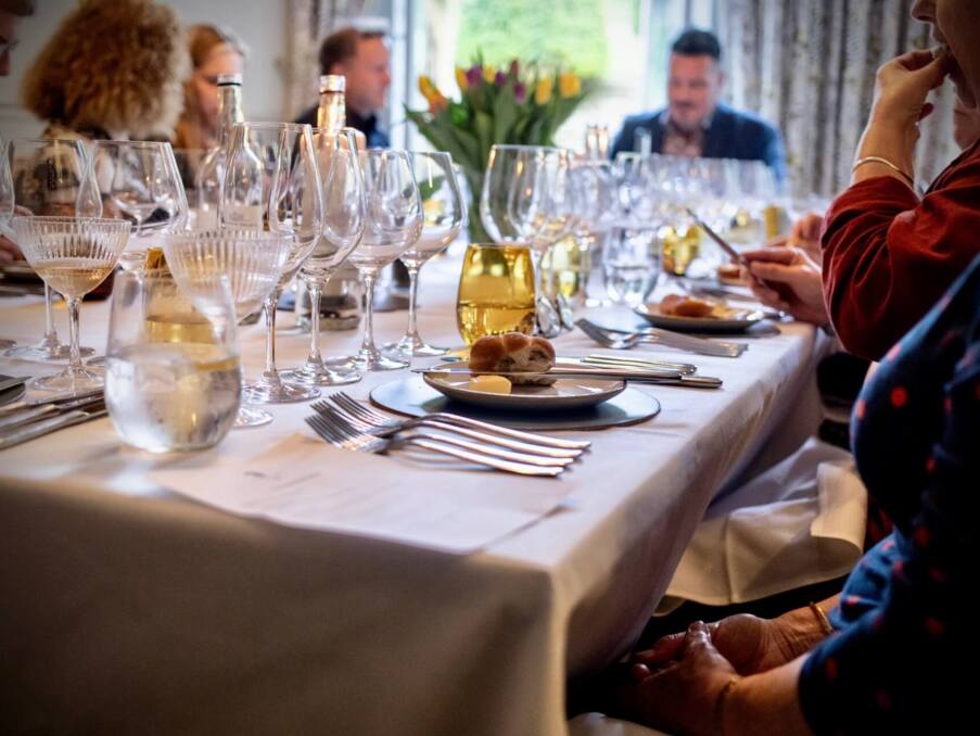 Elegant dining table set with multiple wine glasses, bread rolls, and butter, guests engaged in conversation in a cozy restaurant.