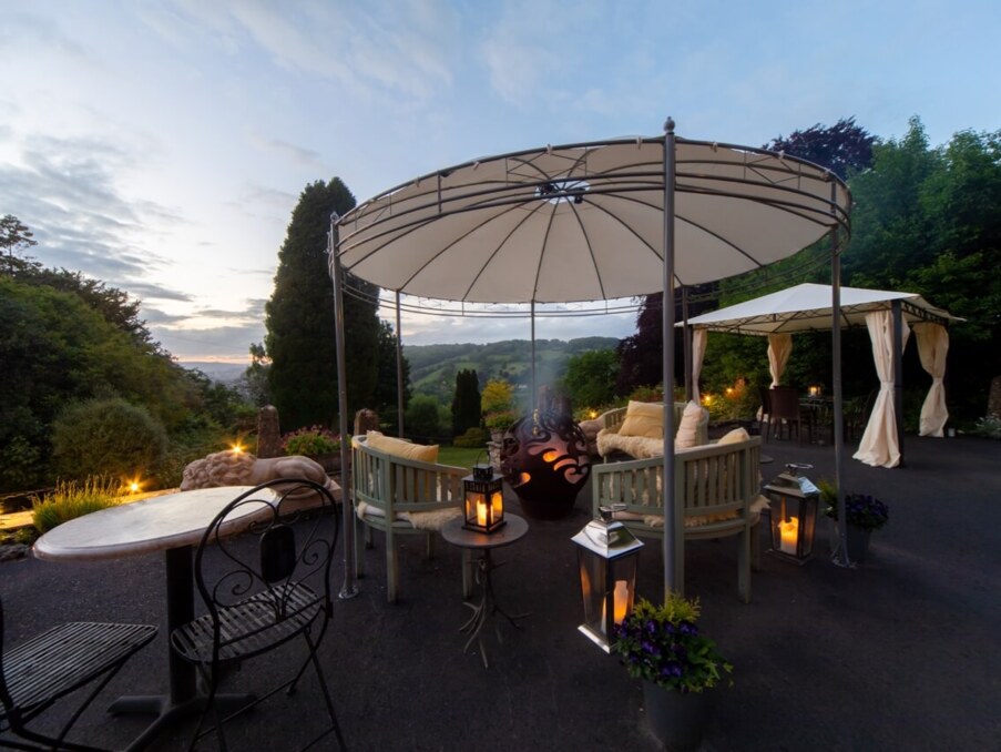 Outdoor seating area with cushioned benches under canopy, lanterns, fire pit, and scenic garden views at dusk