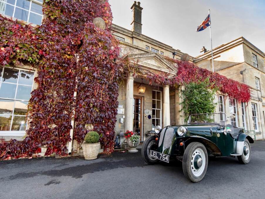 Historic stone hotel entrance with red ivy, classic convertible car parked outside, and British flag fluttering above the building