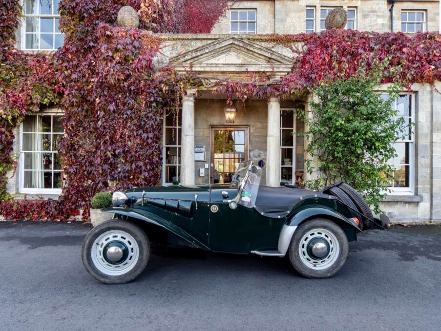 Classic vintage convertible parked in front of a historic stone hotel entrance covered in red ivy with tall columns and large windows
