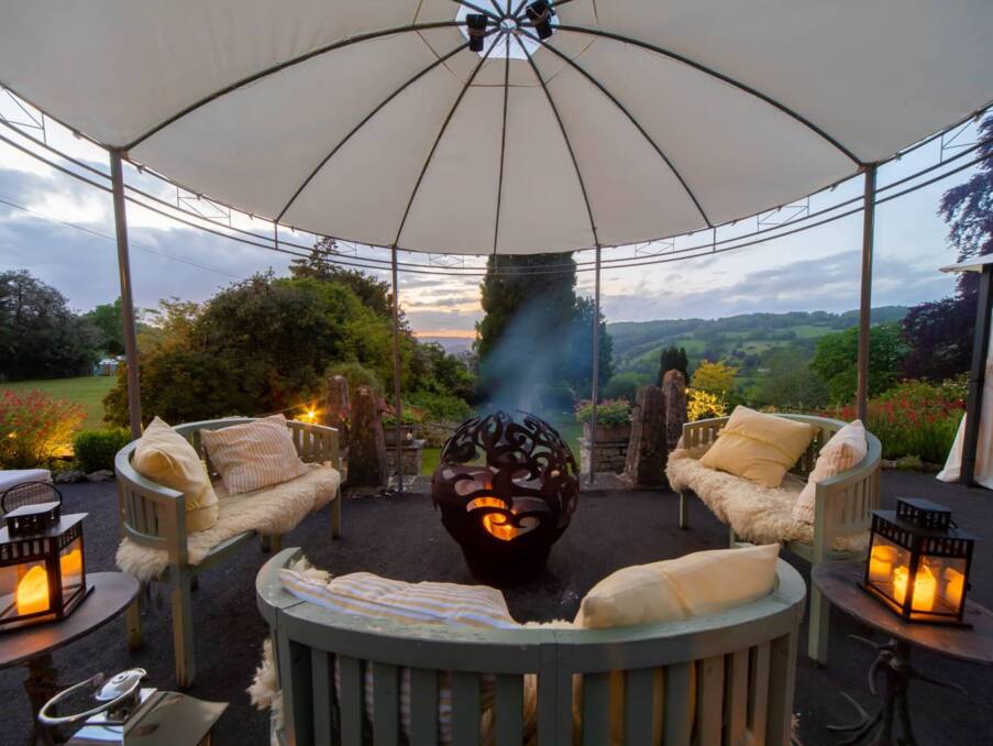 Outdoor lounge area with cushioned benches, decorative fire pit, lanterns, and canopy overlooking a scenic garden and hillside at sunset