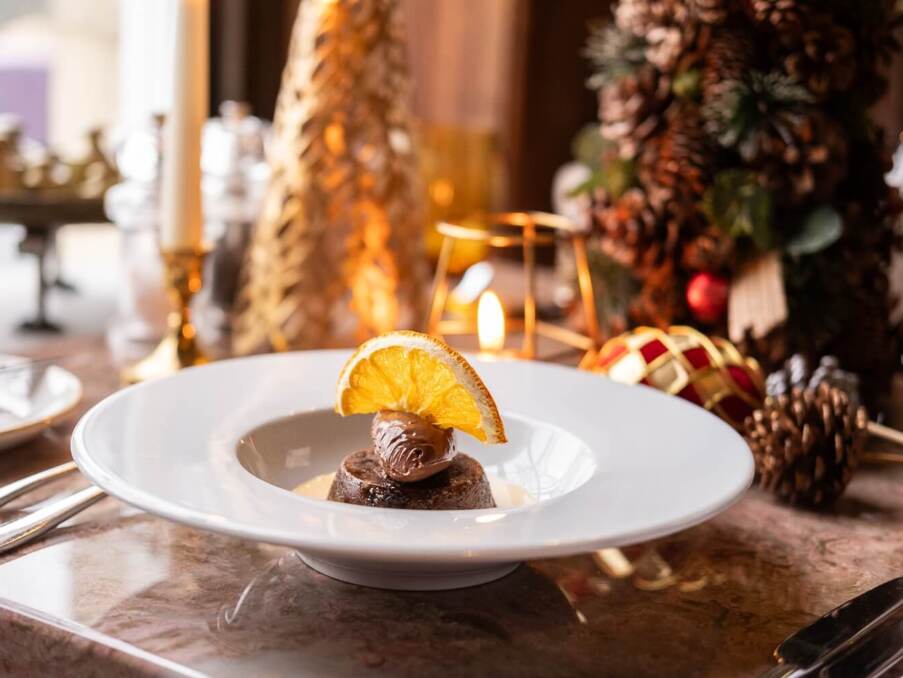Chocolate dessert topped with cream and an orange slice on a white plate, set on a festive table with pinecone decorations and candles