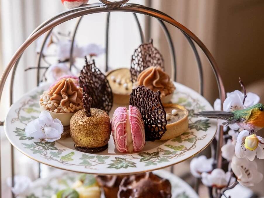Decorative tiered tray with elegant assorted pastries including macarons, cream-topped treats, and a gold-dusted dessert, accented with delicate flowers and a bird