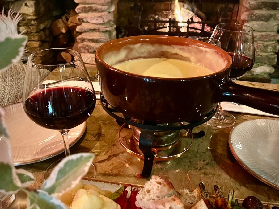Cozy fireplace beside a cheese fondue set with bread, vegetables, and two glasses of red wine on a rustic table