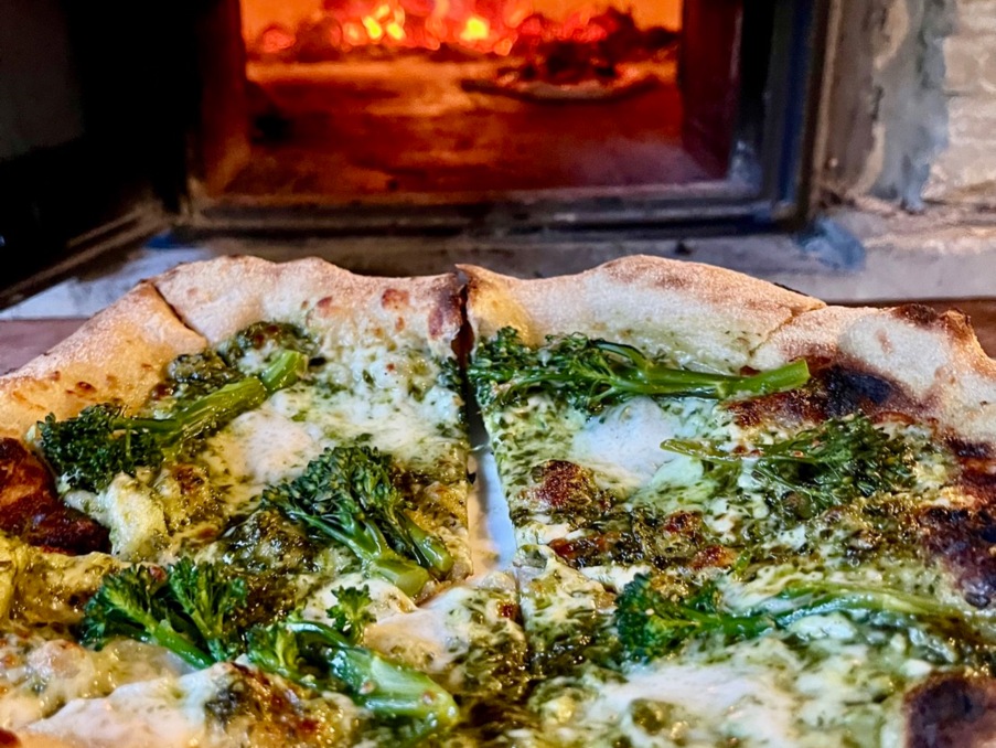 Wood-fired pizza topped with melted cheese and steamed broccoli in front of a rustic stone pizza oven