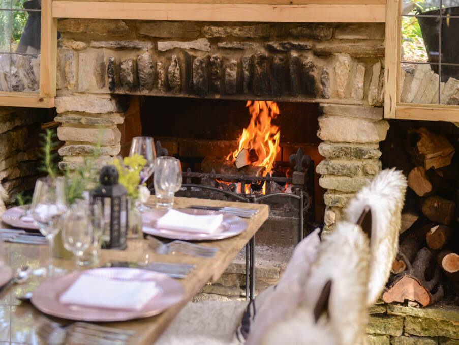 Cozy dining table set with glassware and plates by a rustic stone fireplace with a warm fire and wood stack nearby