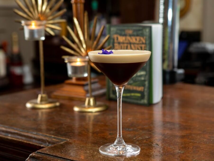 Elegant espresso martini cocktail with creamy foam and purple flower garnish on polished hotel bar counter with candle decor