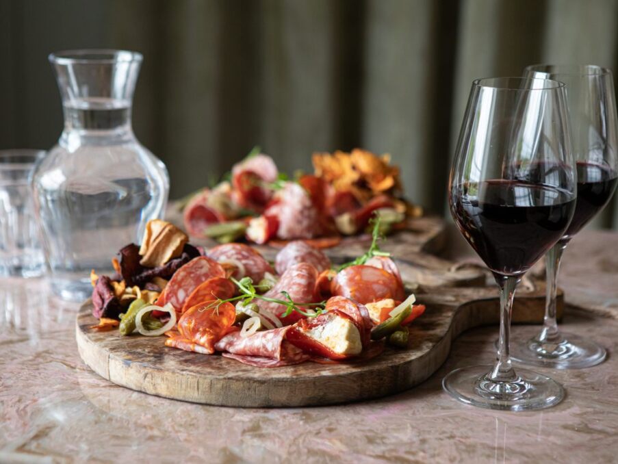 Charcuterie board with assorted cured meats, pickles, and garnishes paired with two glasses of red wine and a carafe of water on a marble table