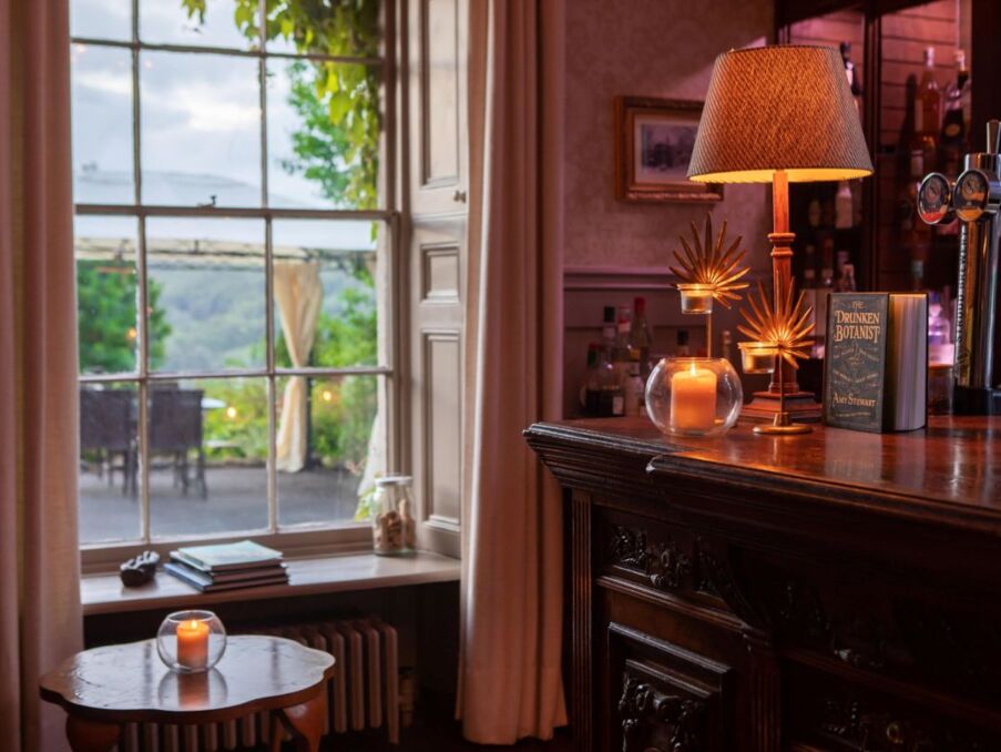 Cozy hotel bar with antique wooden counter, glowing candlelight, decorative lamps, and large window overlooking outdoor seating and greenery