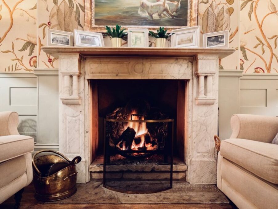Cozy seating area with beige armchairs beside a marble fireplace with a warm fire and framed artwork above