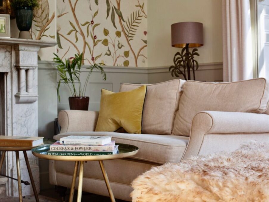 Cozy hotel lounge with beige sofa, decorative yellow pillow, floral wallpaper, marble fireplace, and stack of books on round coffee table