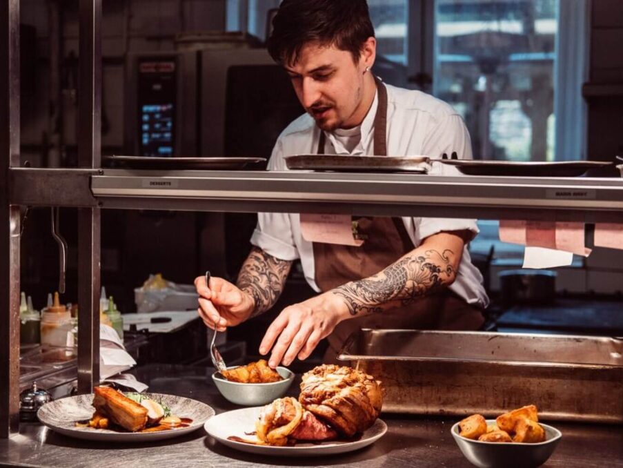 Tattooed chef plating gourmet dishes in professional kitchen, preparing a variety of meats and sides for hotel dining service