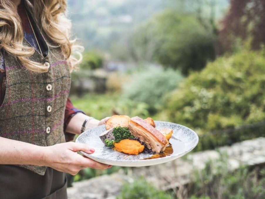 Server holding a plate with roasted pork, mashed sweet potatoes, broccoli, and roasted potatoes in a garden setting