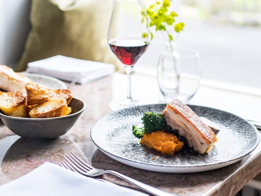 Plate with crispy pork belly, mashed sweet potatoes, and broccoli on a restaurant table with a bowl of roasted potatoes and a glass of red wine