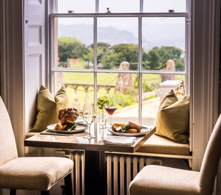 Cozy dining nook with cushioned window seats, two chairs, plates of gourmet food, wine glasses, overlooking lush garden and countryside.