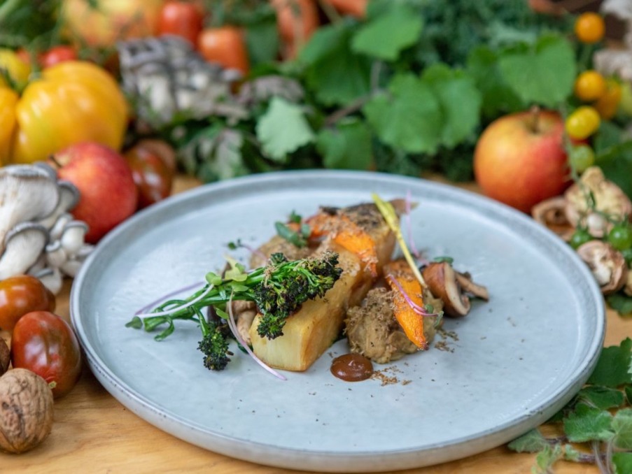 Gourmet vegetable dish with roasted roots, greens, and mushrooms on a ceramic plate surrounded by fresh produce