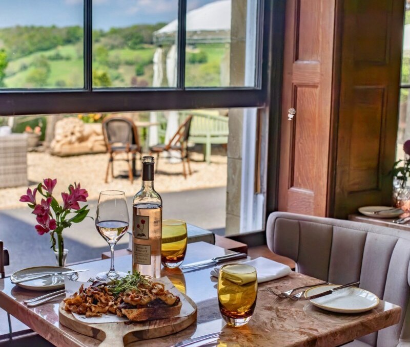 Cozy indoor dining table with wine, gourmet meal on wooden board, fresh flowers, and outdoor countryside view through large window