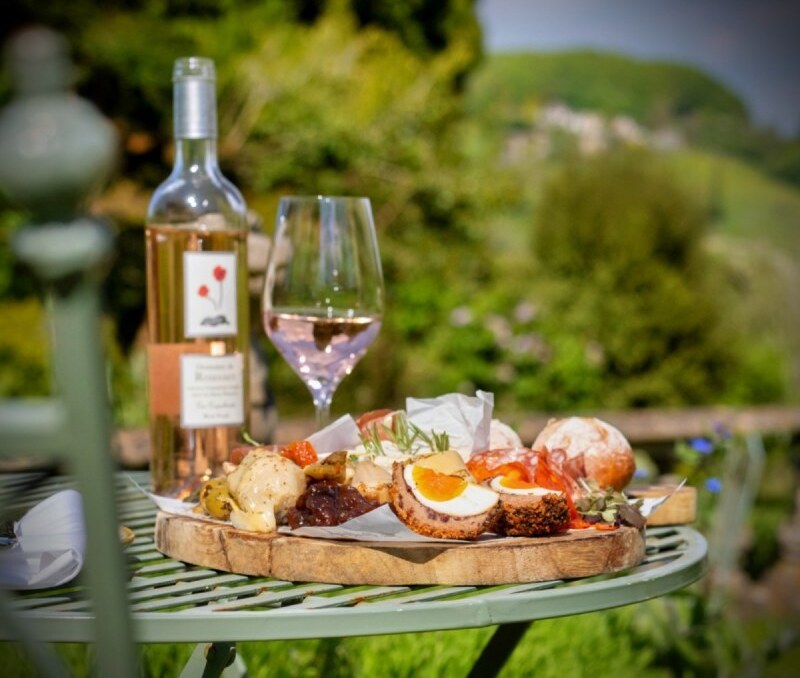 Outdoor table with a wine bottle, glass of rosé, and a rustic wooden board of appetizers including scotch eggs and bread rolls