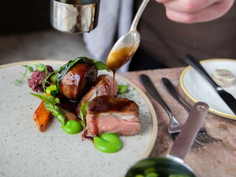 Sauce being drizzled over a plated gourmet dish with tender meat, asparagus, roasted carrot, and vibrant green purée