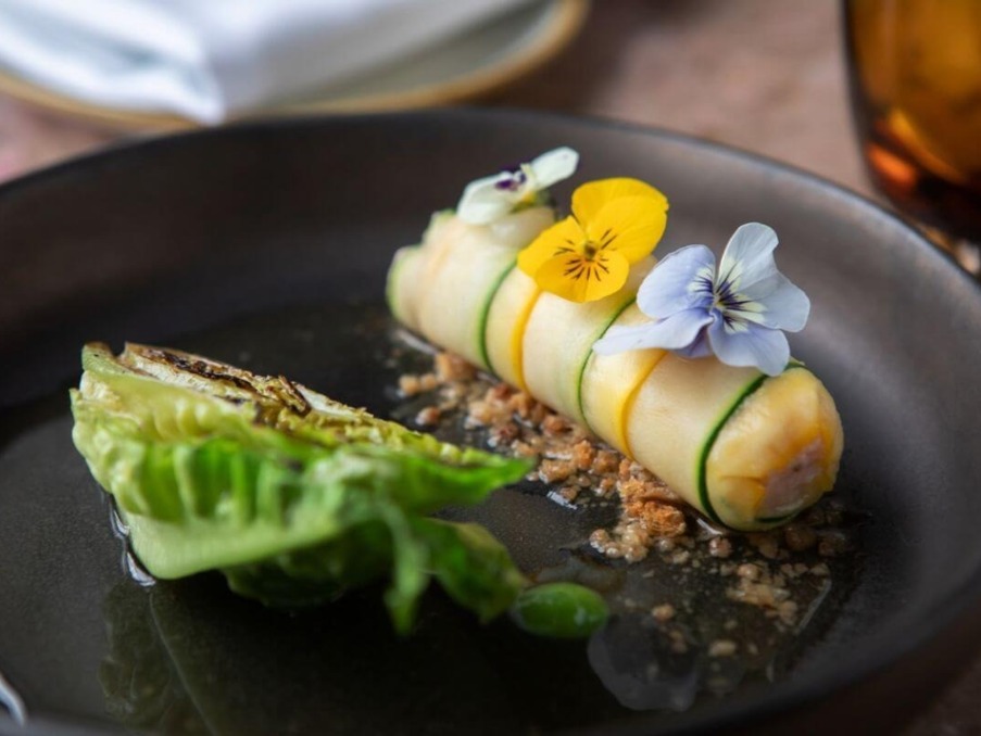 Gourmet dish with rolled vegetable wrapped in thin slices, topped with edible flowers, served with grilled lettuce on black plate