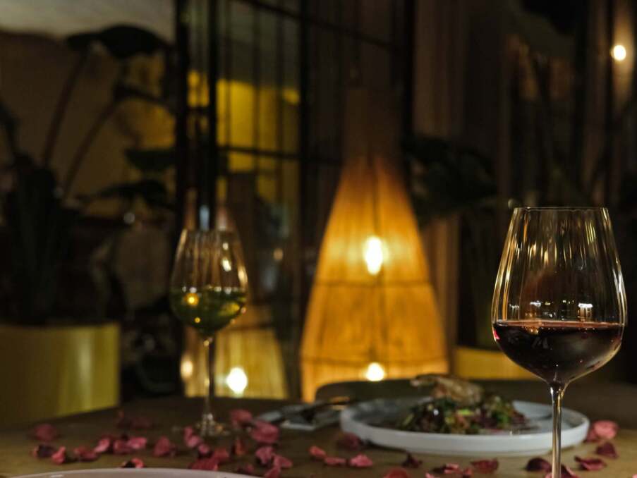 Romantic dimly lit restaurant table with gourmet dishes, red and white wine glasses, and scattered rose petals.
