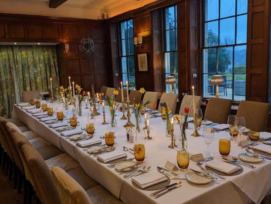 Elegant dining room with long white-clothed table set for a formal dinner, featuring candles, amber glasses, and fresh flowers