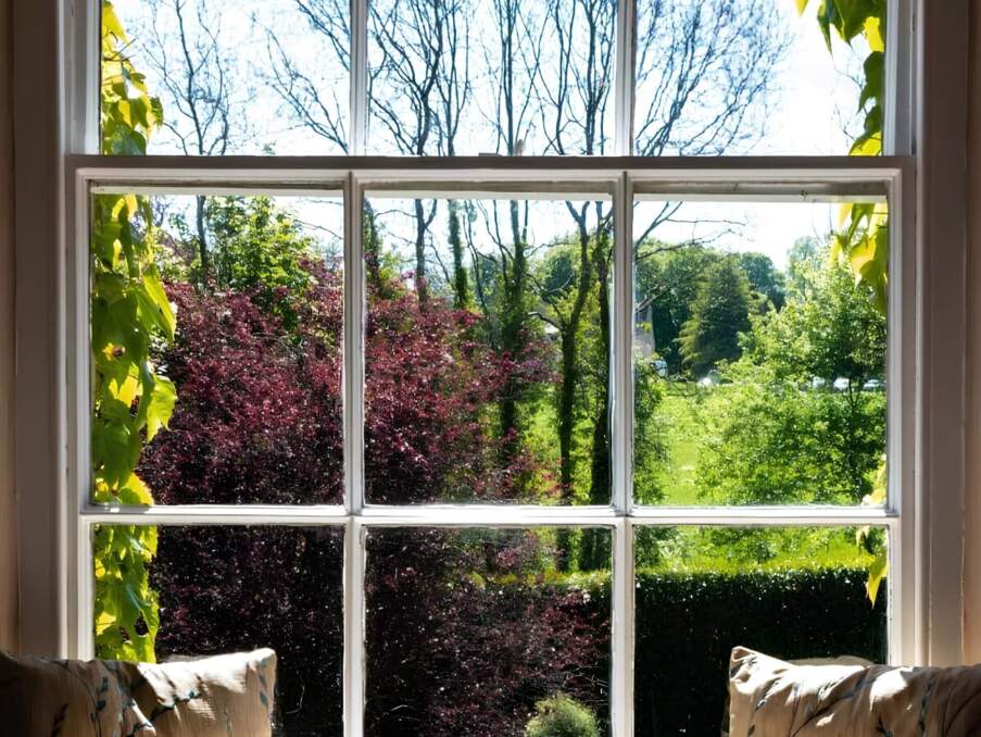 Cozy window seat with cushions overlooking a sunlit garden with lush greenery and colorful trees outside