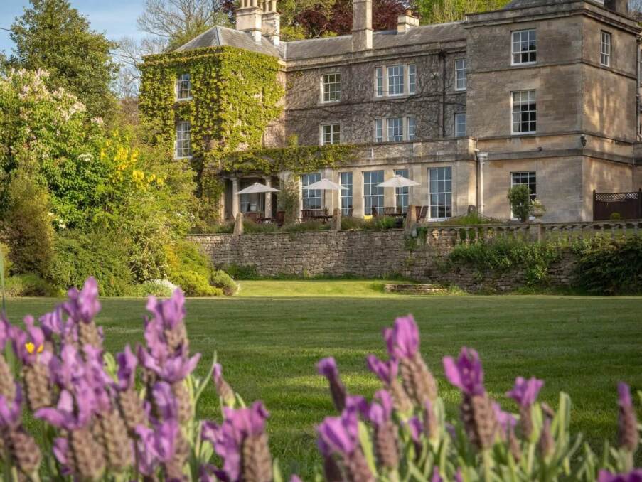 Historic stone hotel with ivy-covered walls, outdoor terrace with umbrellas, surrounded by lush gardens and blooming purple flowers