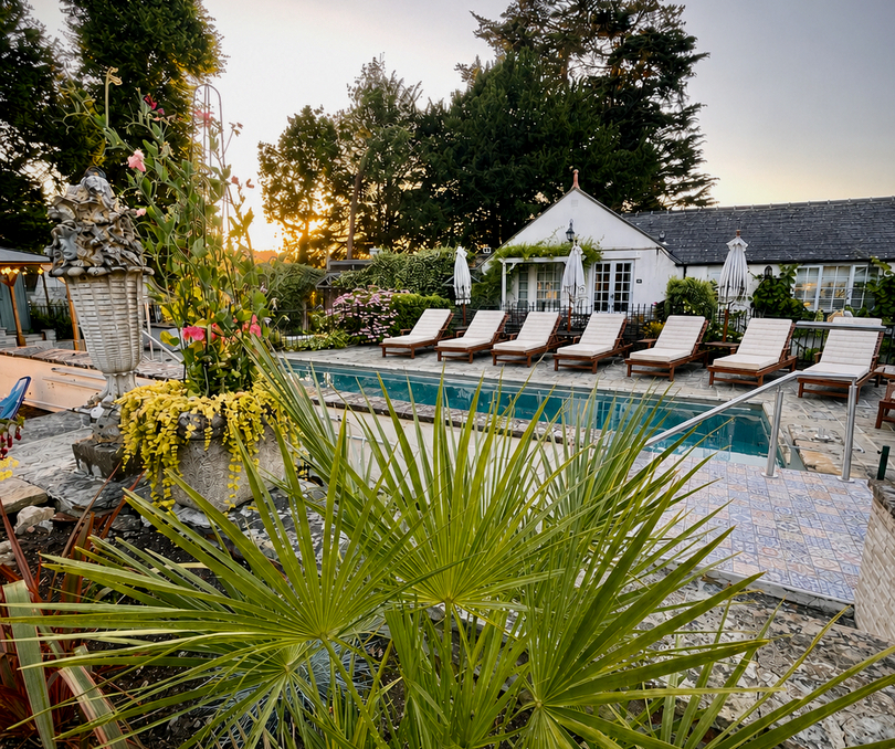 Terrasse de piscine au coucher du soleil avec transats en bois alignés, parasols blancs, petite maison de piscine et palmiers en pots