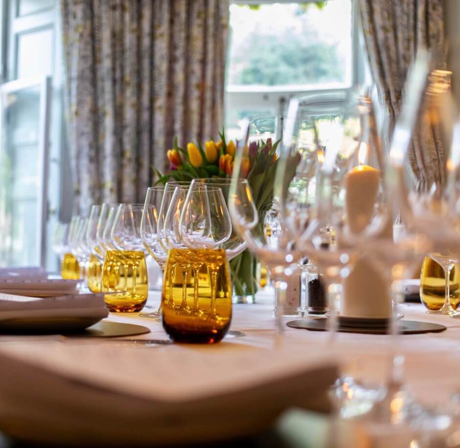 Elegant dining table set with folded napkins, amber glassware, wine glasses, candles, and a floral centerpiece near large windows