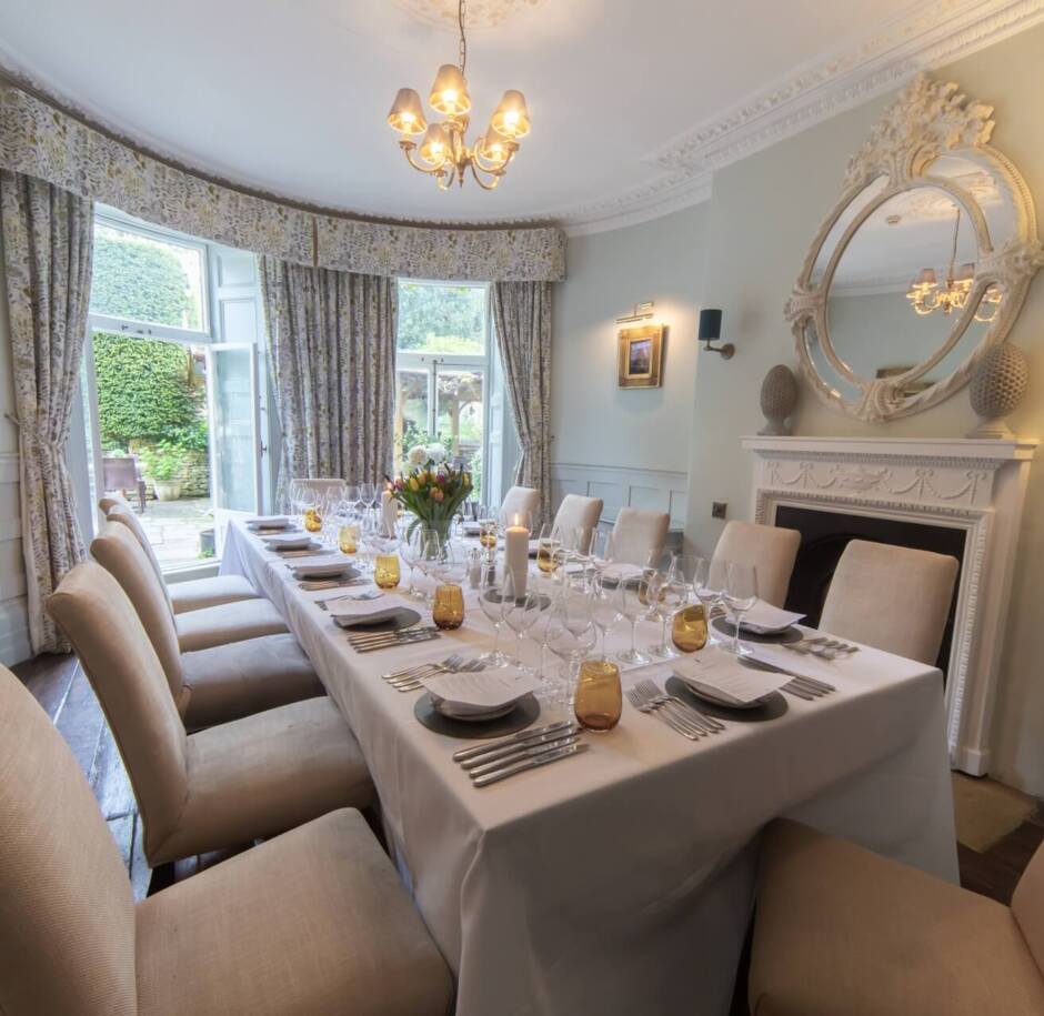 Elegant dining room with ornate fireplace, large round mirror, beige upholstered chairs, floral curtains, and table set for a formal meal