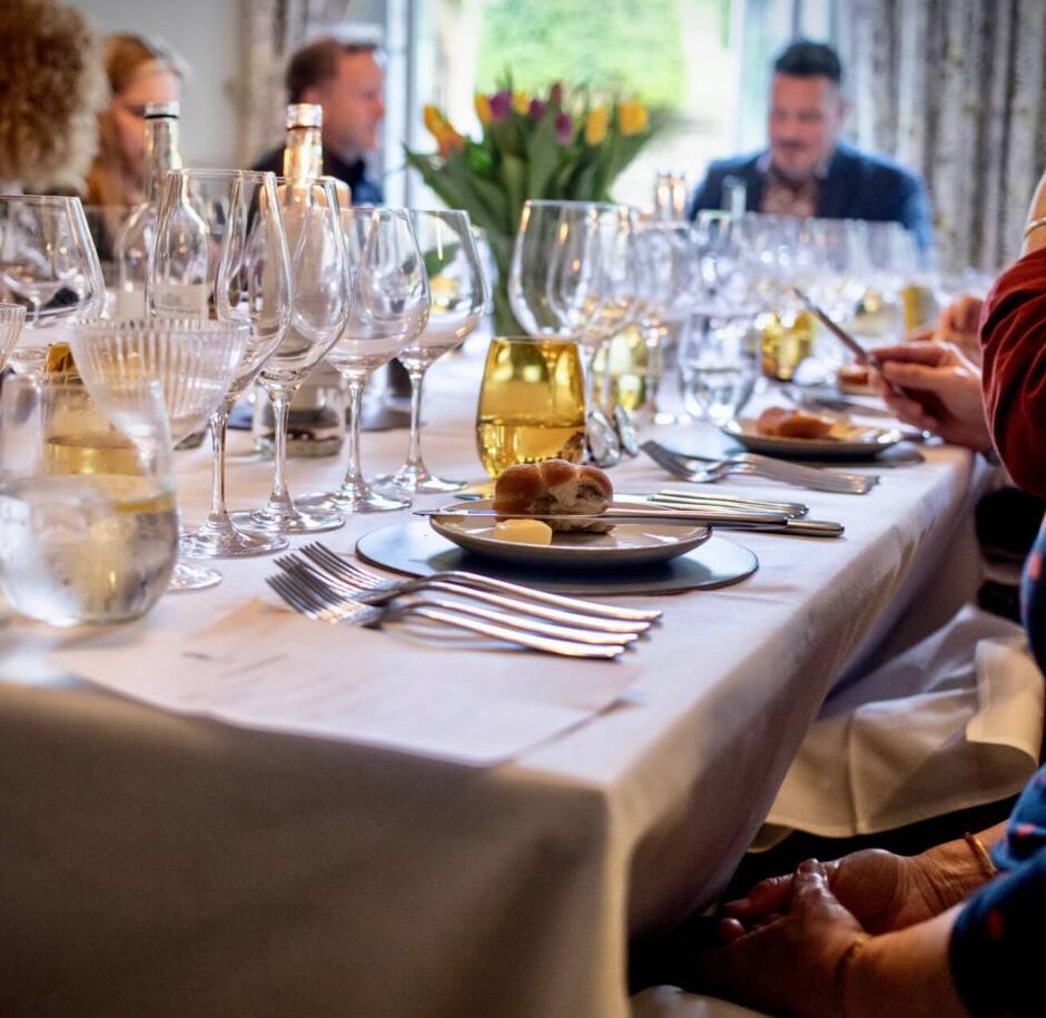 Elegant dining table set with multiple wine glasses, bread rolls, and butter, guests engaged in conversation in a cozy restaurant.