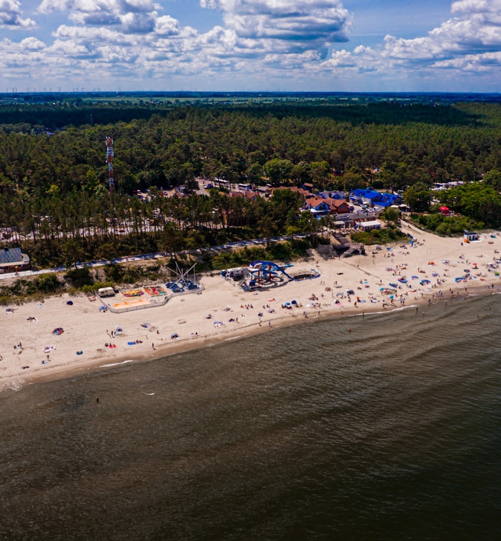 Piaszczysta plaża z atrakcjami wodnymi, parasolami i lasem w tle, idealna na rodzinny wypoczynek nad morzem.