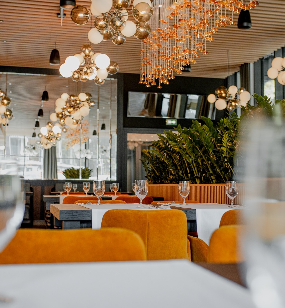 Nowoczesna restauracja z pomarańczowymi krzesłami, białymi obrusami, efektownymi żyrandolami i zielonymi roślinami we wnętrzu